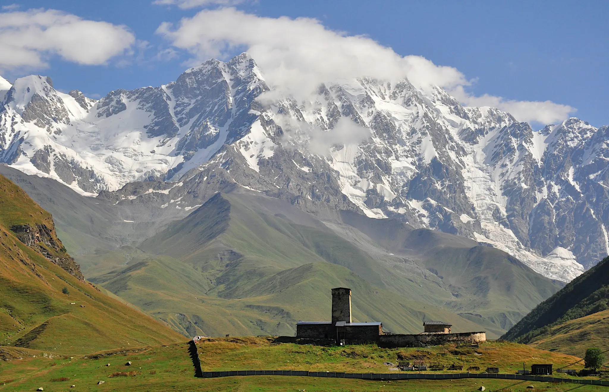 ancient stone tower and buildings in a green valley with snowy mountains